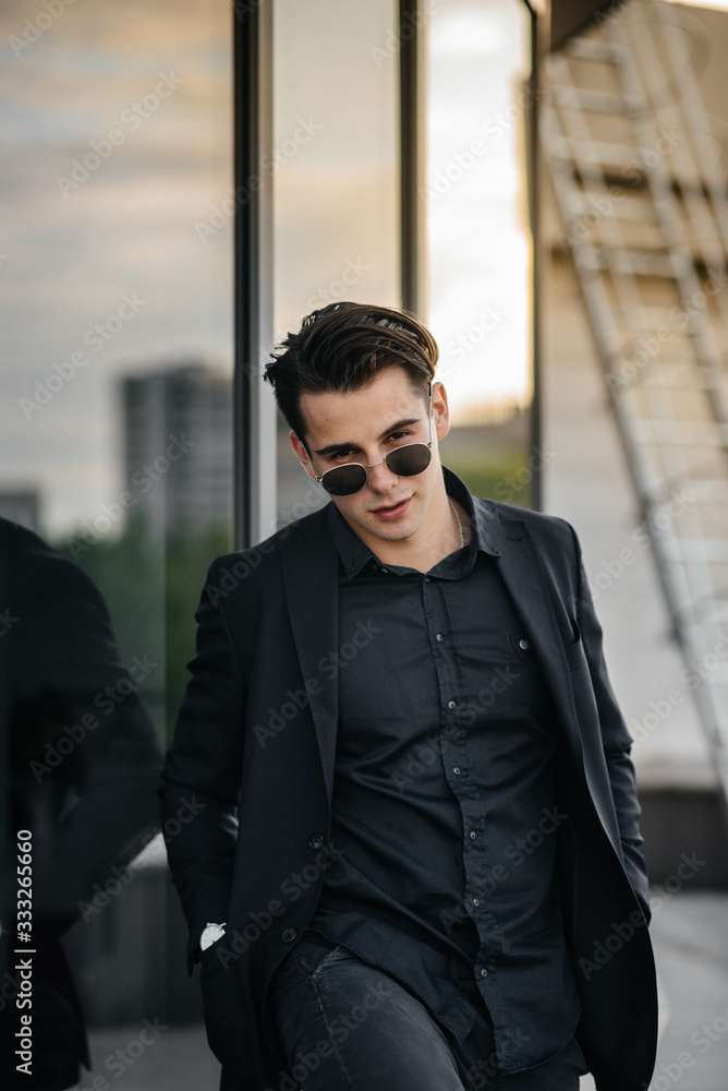 A stylish young man stands near a mirrored building during sunset. Fashion A stylish young man stands near a mirrored building during sunset. Fashion