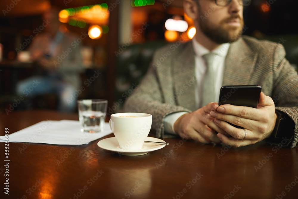 Close-up of young man holding mobile phone while sitting at the table with cup of coffee in cafe