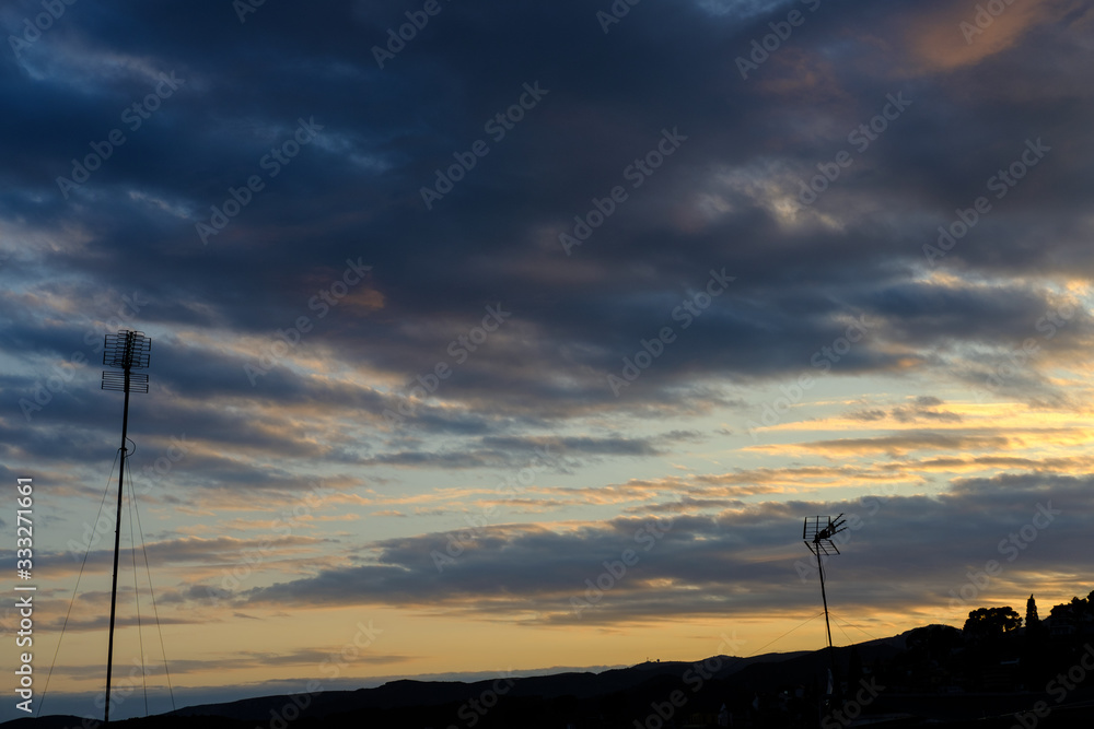 Cloudy sunset over a town with silhouettes of two television antennas.