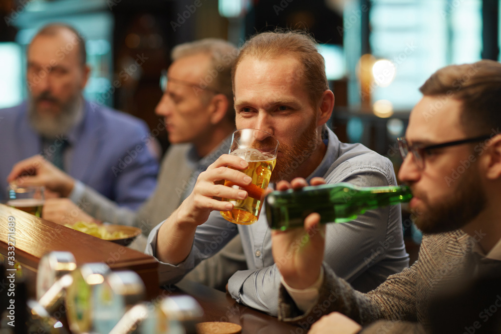 Young man enjoying the glass of beer while sitting at the bar counter together with his friends Young man enjoying the glass of beer while sitting at the bar counter together with his friends
