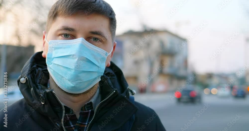 Close up of Caucasian sad ill man in medical mask standing at street and looking to camera. Portrait of guy wearing protection from coronavirus in city. Pandemic concept. Protected person at fresh air Close up of Caucasian sad ill man in medical mask standing at street and looking to camera. Portrait of guy wearing protection from coronavirus in city. Pandemic concept. Protected person at fresh air
