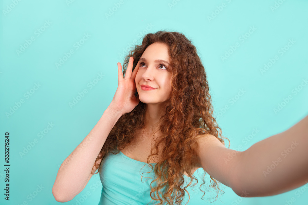 young and beautiful girl with curly hair takes a selfie and smiles on an isolated cyan background young and beautiful girl with curly hair takes a selfie and smiles on an isolated cyan background