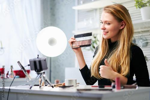 Vlogger female showing eye shadow palette. Beauty blogger woman filming daily make-up routine tutorial near camera on tripod. Influencer girl live streaming cosmetics product review in home studio.