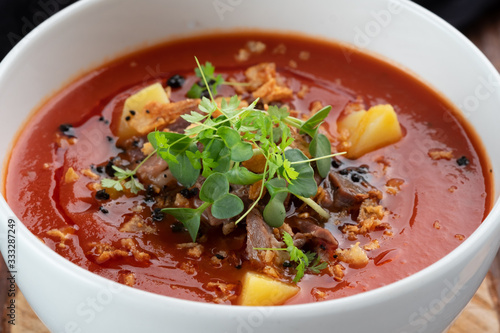 Tomato cream soup with smoked brisket and potatoes in a white bowl on a wooden background, rustic style close-up