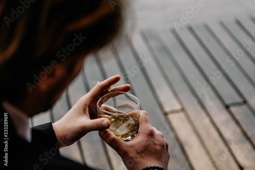 a courageous brutal man dressed in a suit with a glass of strong alcohol and a cigarette in his hands sits in the backyard of his home