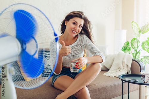 Quadro su tela Happy young woman cooling down by ventilator at home drinking water showing thumb up