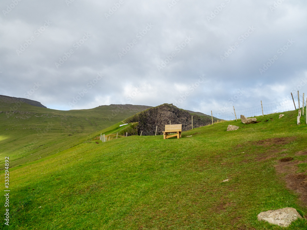 Faroe Islands. Slopes of hills rising above the Gjógv village. Wooden bench in the background. Cloudy sky. Faroe Islands. Slopes of hills rising above the Gjógv village. Wooden bench in the background. Cloudy sky.