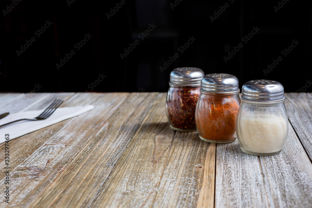 A view of a place setting on a wooden dining table featuring Italian restaurant seasoning, such as parmesan cheese and crushed red pepper. A view of a place setting on a wooden dining table featuring Italian restaurant seasoning, such as parmesan cheese and crushed red pepper.