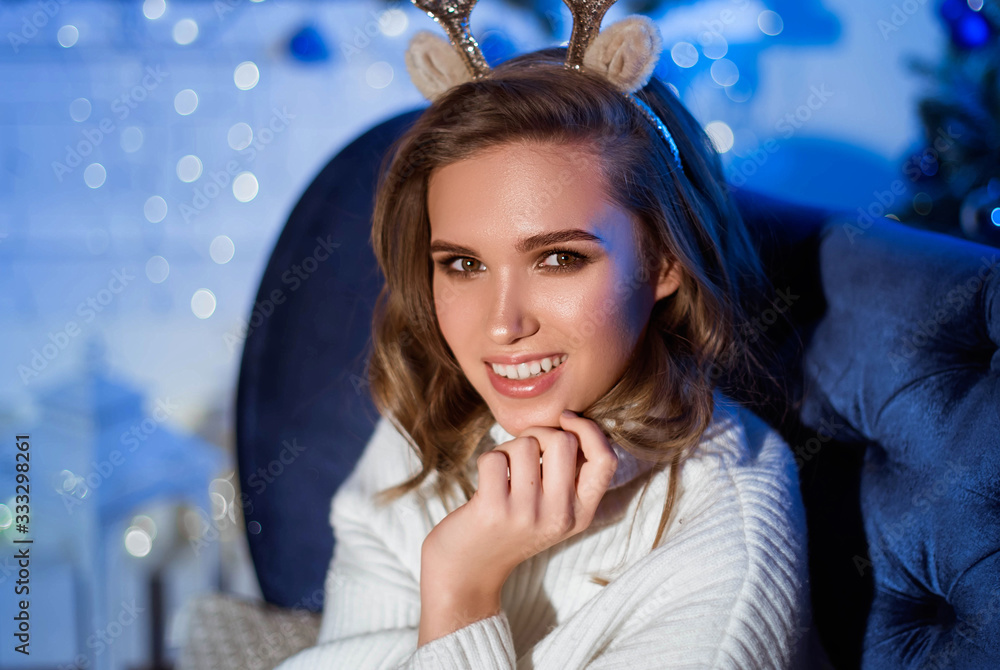 A large portrait of a girl with a broad smile and deer horns on her head sits on a sofa at home. Christmas Eve. New Year.