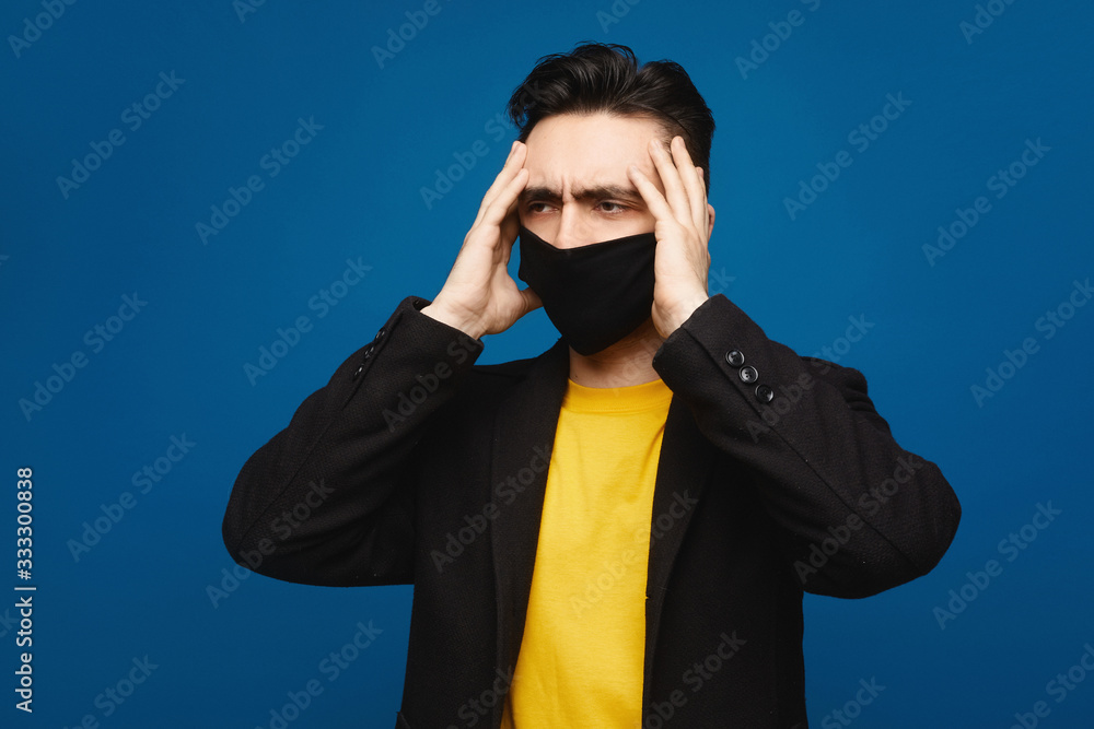 Portrait of young ill man in medical protective mask on face suffering from headache and weakness, isolated at blue background. Typical symptoms of coronavirus COVID-19 or flu. Healthcare concept Portrait of young ill man in medical protective mask on face suffering from headache and weakness, isolated at blue background. Typical symptoms of coronavirus COVID-19 or flu. Healthcare concept