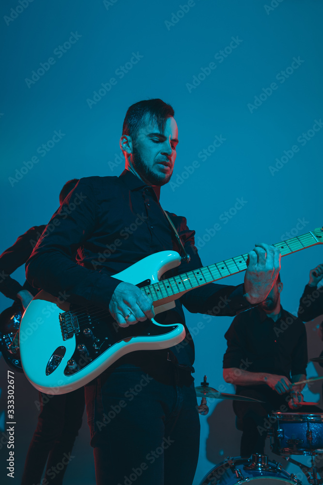 Obraz premium Young caucasian musicians, band performing on blue studio background in neon light. Concept of music, hobby, festival. Colorful portrait of modern artist. Attented and inspired. Art, cover band.