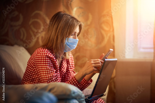 Young european woman wearing surgical face mask stay at home during coronavirus, covid-19 outbreak and looking news or application on smartphone.