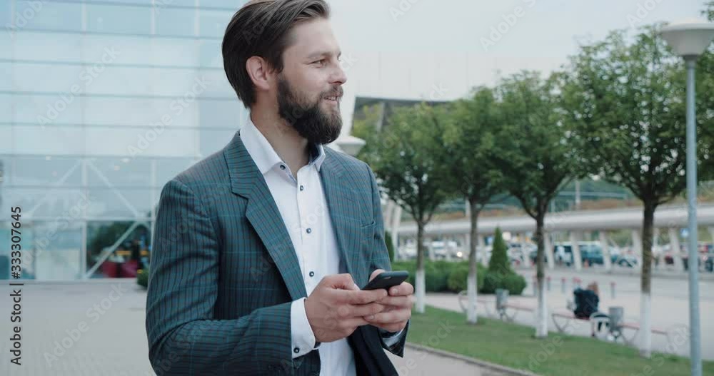 Young Attractive Businessman walking in the City, Using his Smartphone, Looking Stylish. Man in formal suit and eyeglasses. Bearded man with stylish hairstyle having a Walk Outside Officeю Young Attractive Businessman walking in the City, Using his Smartphone, Looking Stylish. Man in formal suit and eyeglasses. Bearded man with stylish hairstyle having a Walk Outside Officeю