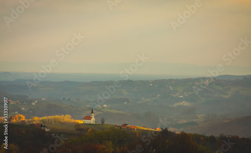 Lonely church in autumn setting in the region of Bela Krajina or White Carniola in Slovenia on a sunny hazy day. Visible lovely hills and houses on top of them. Romantic autumn setting