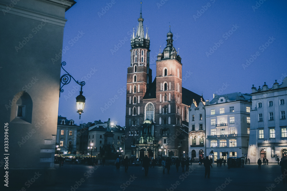 Naklejka premium Mariacka bazylika or St. Mary Basilica in Krakow, Poland in beautiful night setting and blue light. Visible red brick towers and blue sky with some tourists around