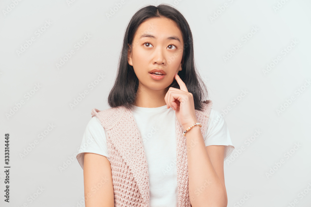 Girl looks up thoughtfully, holding index finger to cheek or chin. young Asian appearance with black hair and brown eyes dressed in t-shirt and pink cardigan stands isolated white background Studio Girl looks up thoughtfully, holding index finger to cheek or chin. young Asian appearance with black hair and brown eyes dressed in t-shirt and pink cardigan stands isolated white background Studio