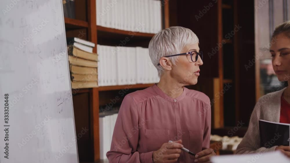 Professor helping student in library at college. Group of multiethnic students taking an active part in a lesson while standing in library. Mature woman teacher and university student discussing. Professor helping student in library at college. Group of multiethnic students taking an active part in a lesson while standing in library. Mature woman teacher and university student discussing.