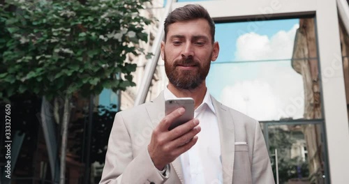 Sucessfull Businessman browsing his Smartphone near modern Office Building. Attractive Man is smiling and wearing elegant Suit Style. Luxury Lifestyle. Successfull Business Man in Crisis. photo