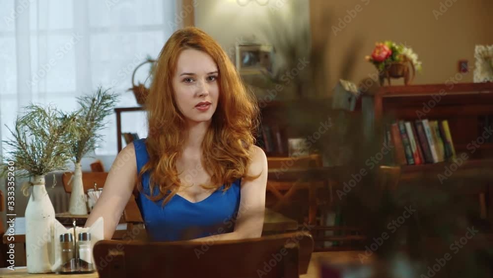The beautiful sexy girl with long red hair in blue dress sitting in cafe and smiling. Slowmo. The beautiful sexy girl with long red hair in blue dress sitting in cafe and smiling. Slowmo.