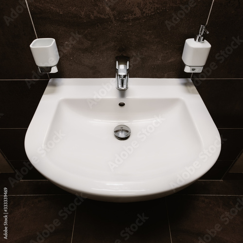 Medium-size U-shaped washbasin in white.Completed with chrome,lever,faucet and plug.There is bathroom accessories like soap dispenser and ceramic cup for brushes.In background are dark colored tiles.