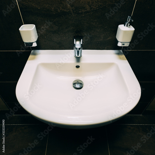 Medium-size U-shaped washbasin in white.Completed with chrome,lever,faucet and plug.There is bathroom accessories like soap dispenser and ceramic cup for brushes.In background are dark colored tiles.