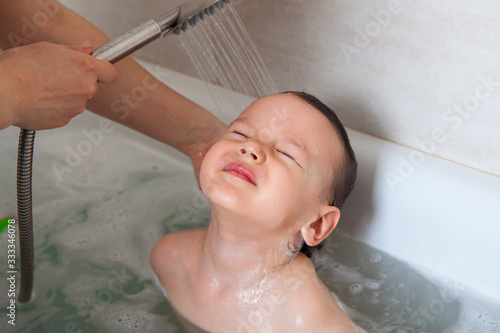 Mom washes her baby's head in a bath with water. Flushing shower shampoo foam. Shampooing without tears.