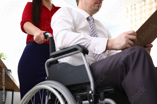 Close-up of woman co-worker accompany man in disabled carriage. Employee reading important papers. Business and adaptation of people with disabilities in society concept