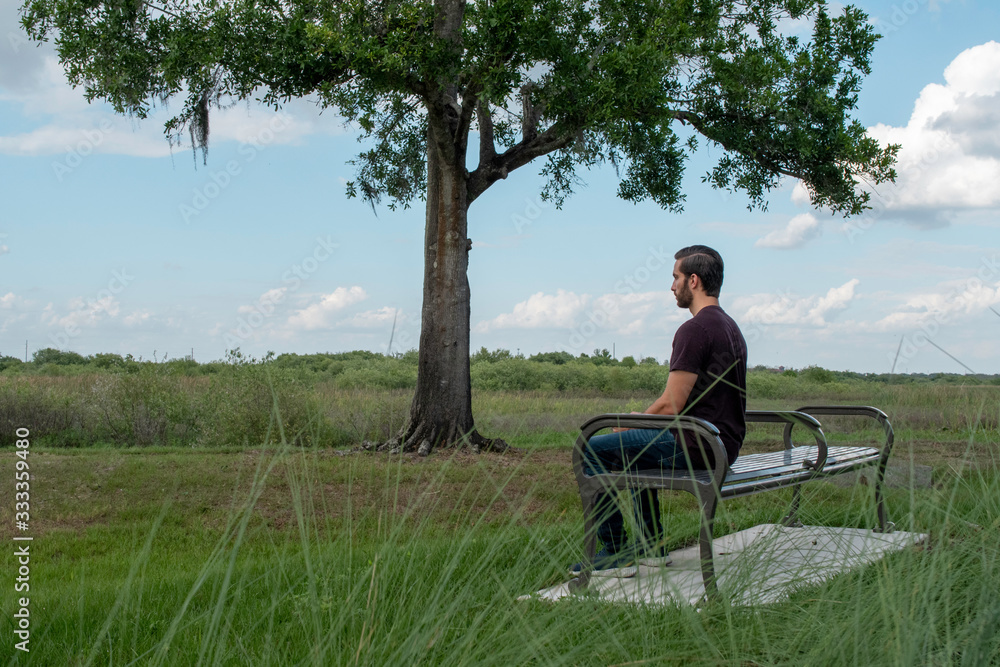 Fototapeta premium A man with a red shirt and blue jeans relaxes on a park bench playing with his phone and admiring the world around him on a bright sunny day on a park bench
