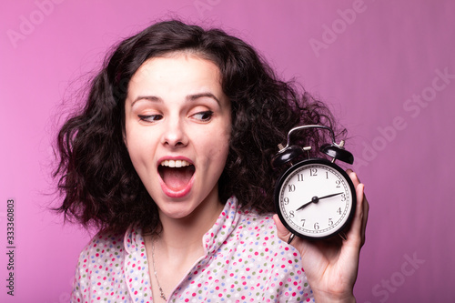  beautiful curly girl in pajamas holds alarm clock in hands, pink background