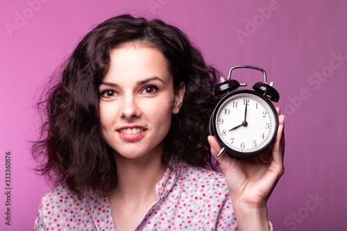  beautiful curly girl in pajamas holds alarm clock in hands, pink background