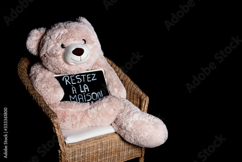 Giant fluffy teddy bear sitting on a wood chair with his message 'Restez à la maison' on a blackboard