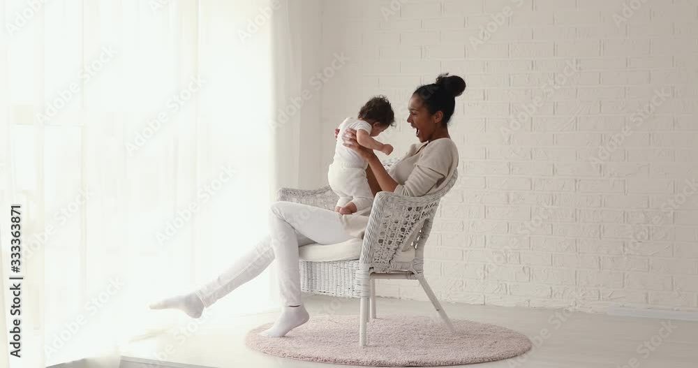 Cheerful young african mum lifting cute infant son or daughter up having fun in cozy white room. Happy single mother holding baby girl playing enjoying sweet moments of love sitting on chair at home. Cheerful young african mum lifting cute infant son or daughter up having fun in cozy white room. Happy single mother holding baby girl playing enjoying sweet moments of love sitting on chair at home.