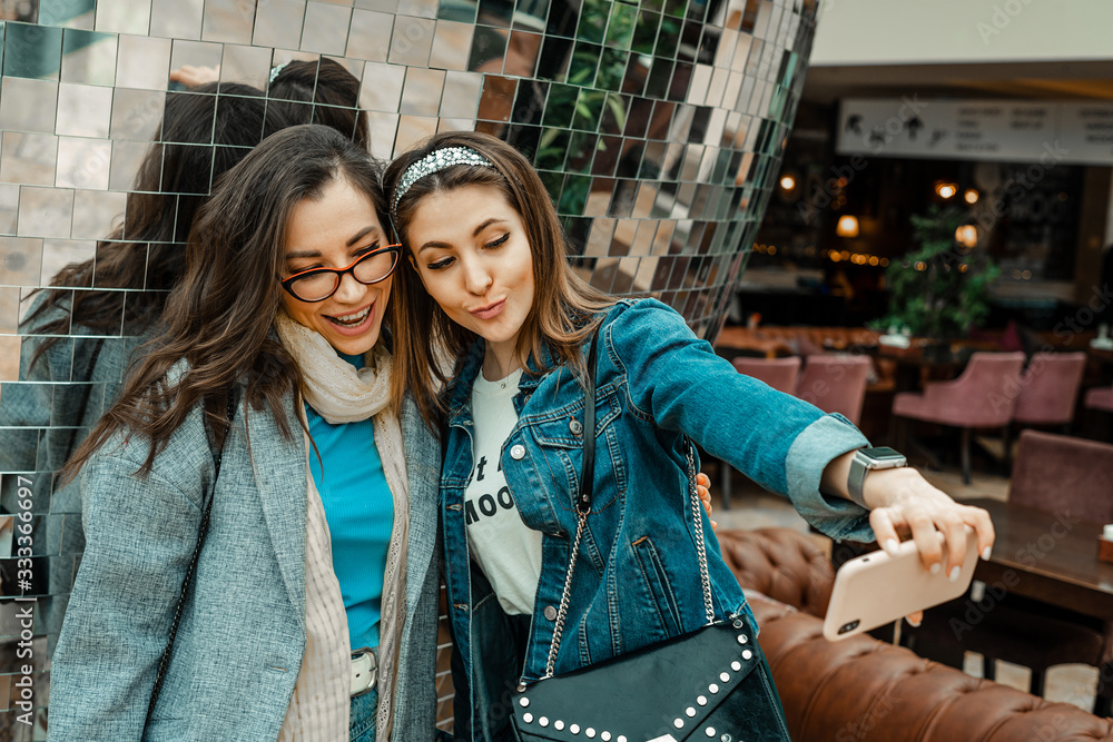 Fototapeta premium Trade, buyers. Two beautiful girls make purchases in a shopping center, go shopping. They look at the smartphone, take a selfie. The joy of consumption.
