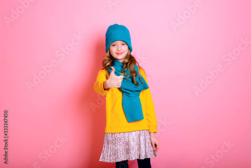 Blonde kid child girl is dressed in a warm sweater, scarf and beanie on a pink background. Fight the cold. Winter and autumn seasons. Concept of children's health, vitamins, medicines.