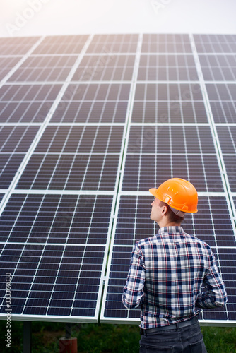 Engineer in an orange helmet stands with his back to the camera against the background of a large plantation of solar panels. Home construction. Green ecological power energy generation.