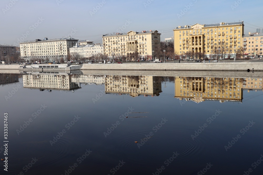 Buildings of the Frunze Embankment in Moscow are reflected in the smooth blue water of the Moscow River. Early spring, sunny day.