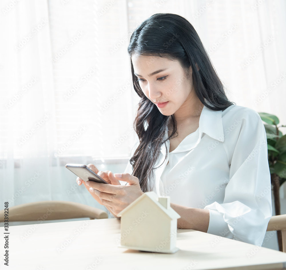 Young beauty Asian business woman in casual hand using smart mobile phone sit at home with house model, planing to buy or rent home concept