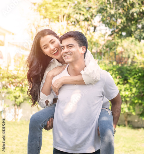Portrait of cute romantic caucasian guy in checkered shirt, dreamy asian lady rides him on rear. Leisure, chill happiness, lawn stroll, relax, romance lifestyle, well dressed partners posing © mkitina4