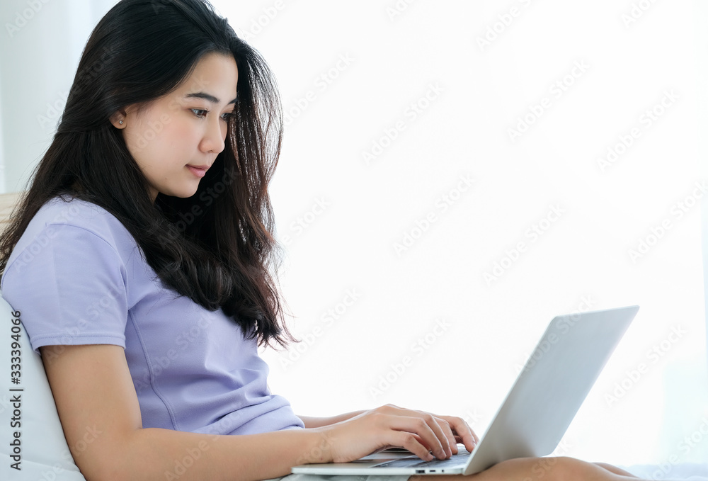 Pretty Asian woman sitting on the bed and using laptop to see social media and do shopping online isolate on white background. Feeling happy and relaxation. Working at home and technology concept.