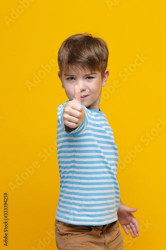 Little cute boy in striped clothes shows a thumb raised up on a hand outstretched in front isolated on a yellow background.