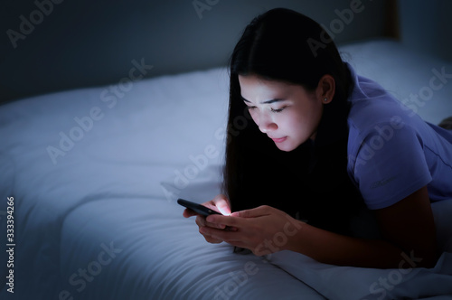 Asian young woman lying on the white bed and playing smartphones during night time. She is chatting with her friend. Using phone in low light is impact to eyes. Health and social Concept
