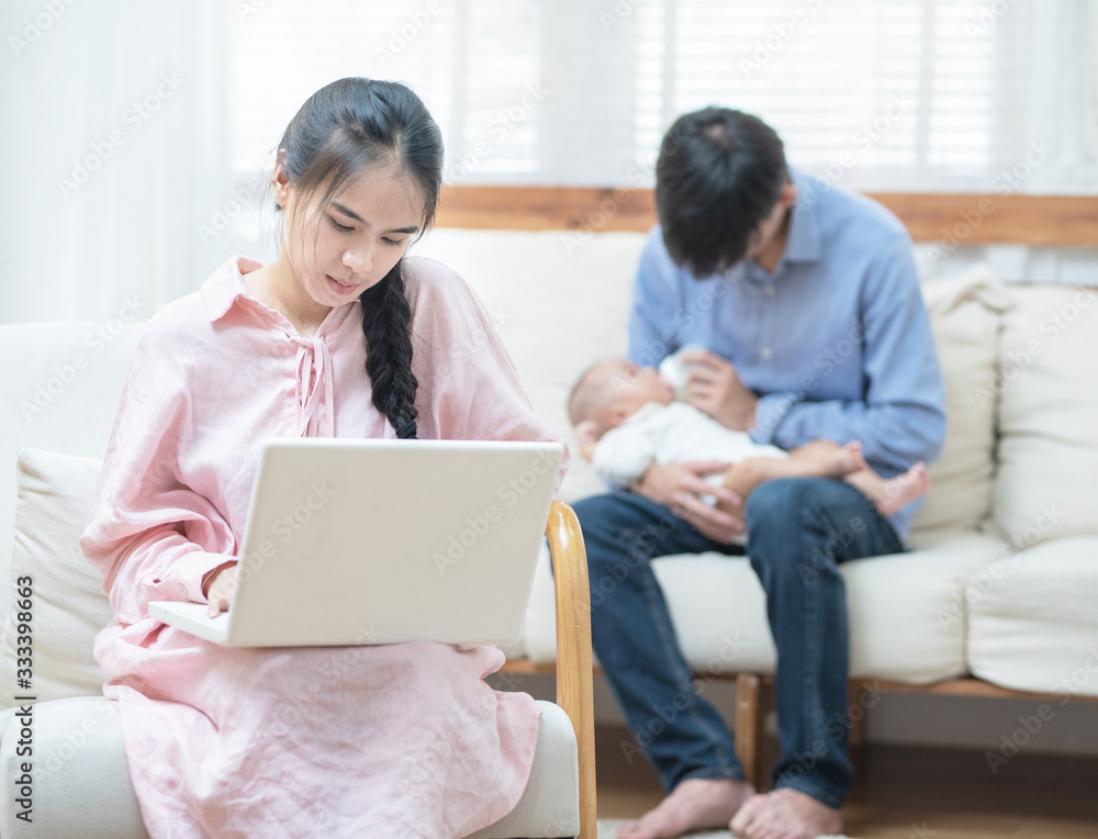 Young asian mother using laptop computer in living room at home with blurred of father acting mom feeding milk bottle in background his son baby 1 year old, work at home and social distancing concept. Young asian mother using laptop computer in living room at home with blurred of father acting mom feeding milk bottle in background his son baby 1 year old, work at home and social distancing concept.