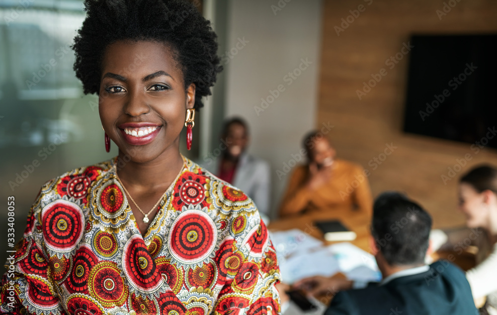 © JonoErasmus - close up of beautiful young smiling professional black african business woman, coworkers hold a meeting in background