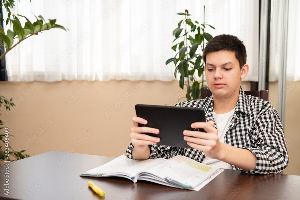 Caucasian teen boy learning language online using digital tablet looking at screen, doing school tasks at home, writing notes, listening lecture. E-learning and distance education concept.