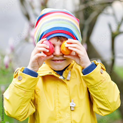 Lovely adorable little kid boy making an egg hunt on Easter. Happy child searching and finding colorful eggs in domestic garden. Boy in spring clothes on cold day. Old christian and catholoc tradition photo