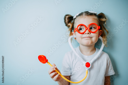 Baby girl in white shirt holds stethoscope. A kid doctor shows how to act while pandemic of covid-19.