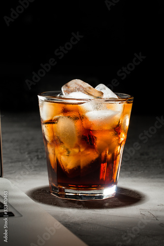 glass of cola with ice and laptop on grey stone table on black background