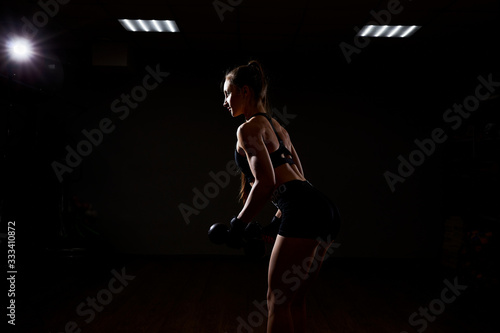 Young beautiful girl athlete bodybuilder does exercises on dark background. Concept - the power of beauty, diet, sports.