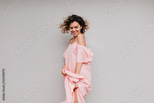 Graceful tanned woman playing with her long pink dress. Indoor shot of charming caucasian lady with wavy hairstyle.