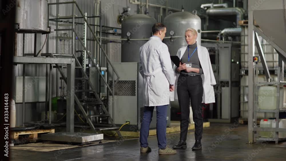 Full length wide shot of middle aged female inspector with clipboard and id card talking to chief engineer standing in factory, both wearing white coats Full length wide shot of middle aged female inspector with clipboard and id card talking to chief engineer standing in factory, both wearing white coats